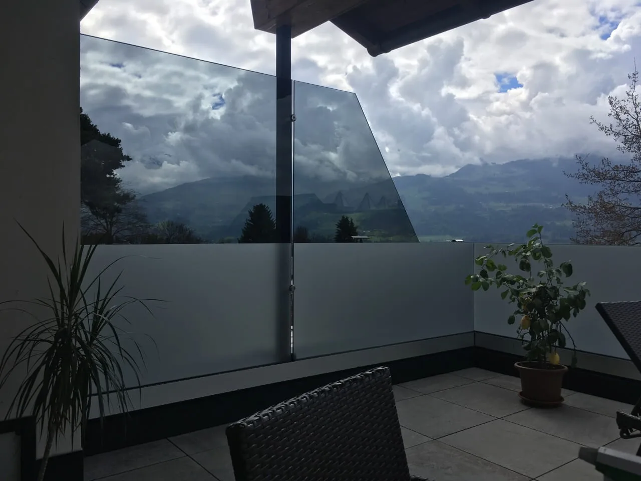 Glasgeländer auf einer Terrasse, dahinter Bäume und Berge
